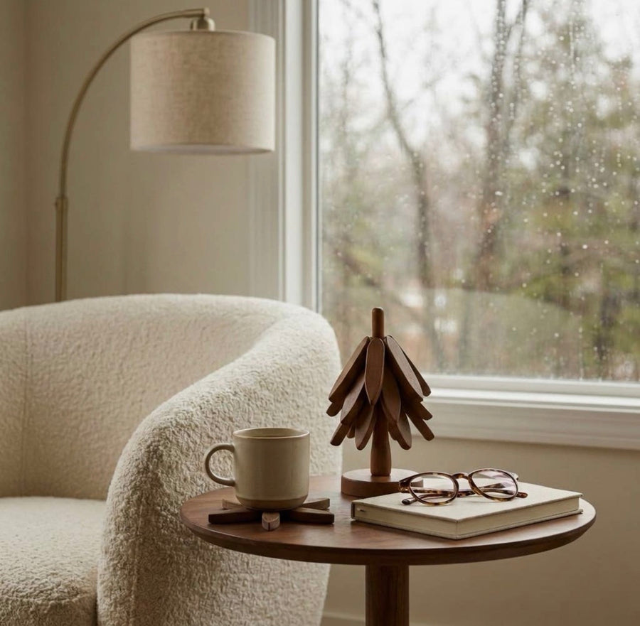 The Grove in Evening on a round side table by a rain-streaked window, beside a mug, reading glasses and a book.