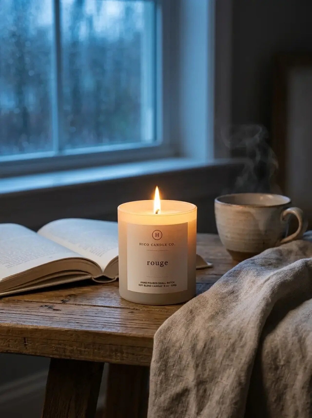 Hico Rouge candle on a wooden table by a window with a book and mug.